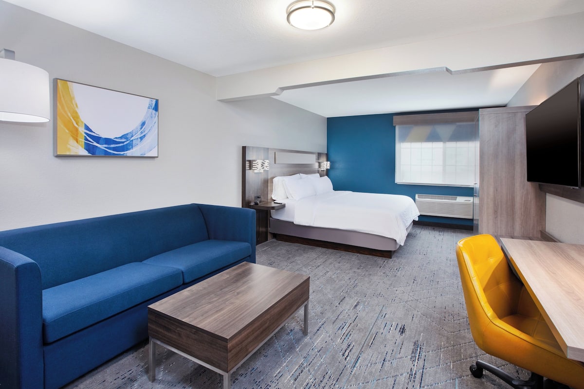 A hotel room with a bed, a blue sofa, a wooden coffee table, a yellow chair, a flat-screen TV, and a painting on the wall.