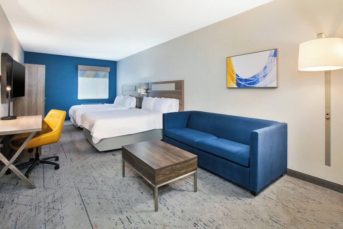 A hotel room with a bed, a blue sofa, a wooden coffee table, a desk with a chair, and a TV.
