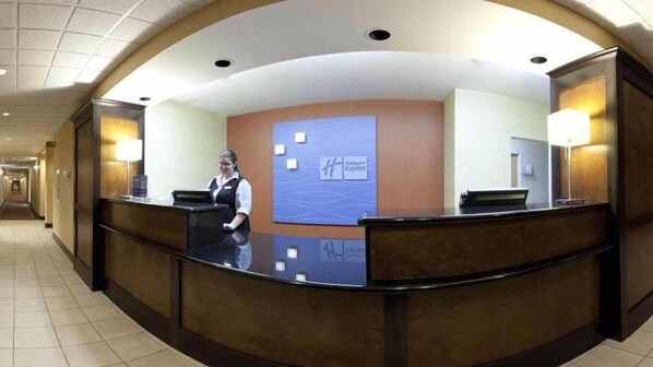 A reception desk with a receptionist, a blue wall with a logo, and a hallway with doors. - Holiday Inn Express & Suites Wauseon by IHG (Wauseon)