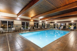 Une piscine intérieure dotée d’un sol carrelé, de poutres en bois au plafond et de plusieurs espaces de détente avec des chaises et des tables.