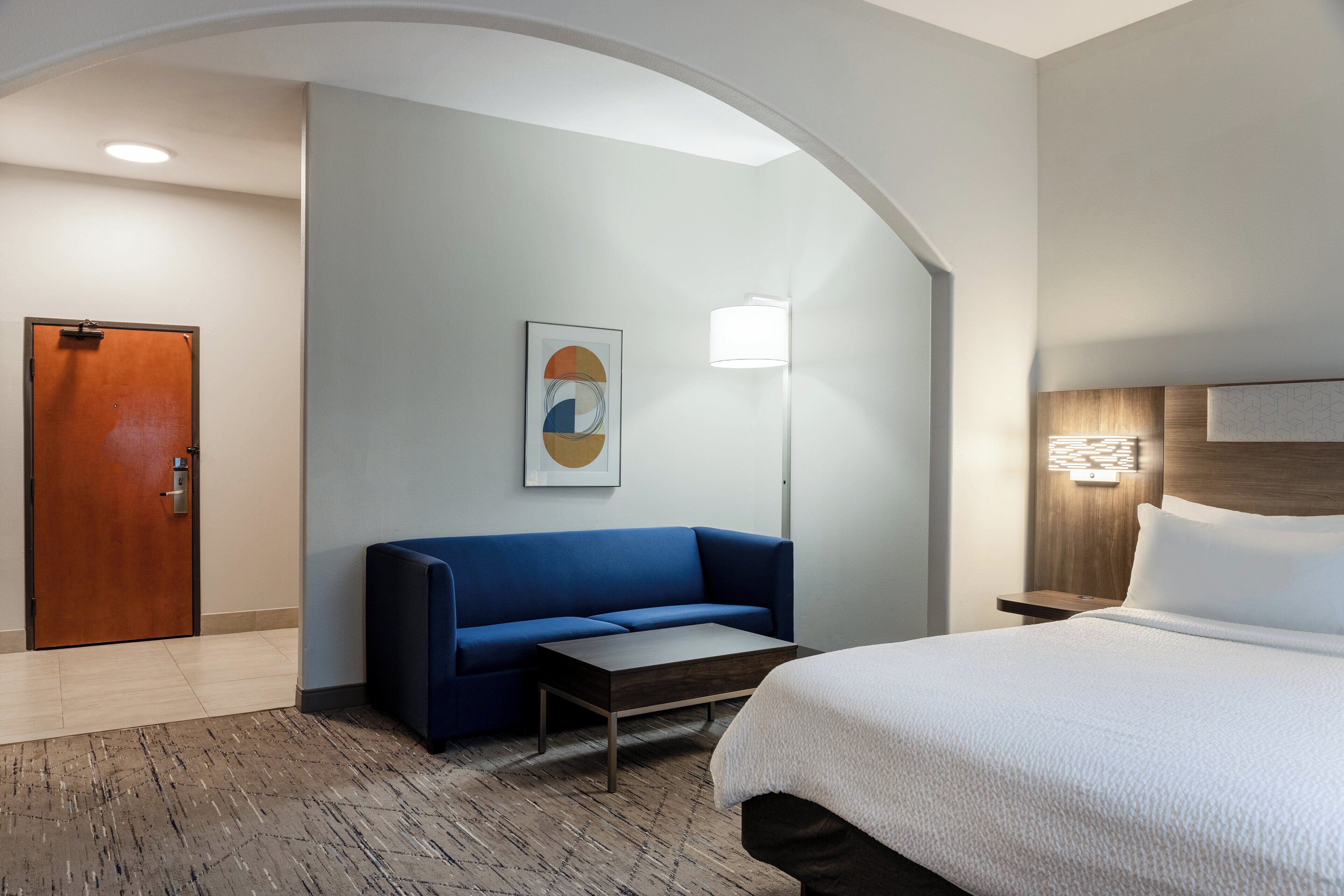 Une chambre d’hôtel avec un lit, un canapé bleu, une petite table, une lampe et une porte.