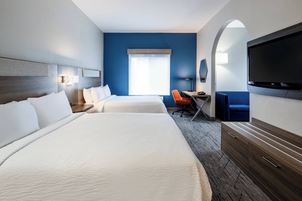 Ein Hotelzimmer mit zwei Betten, einem Schreibtisch, einem orangefarbenen Sessel, einem Fernseher und einem großen Fenster.