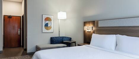 Una habitación de hotel con una cama grande, una zona de estar con una silla y una mesa, una lámpara fijada a la pared y una puerta.