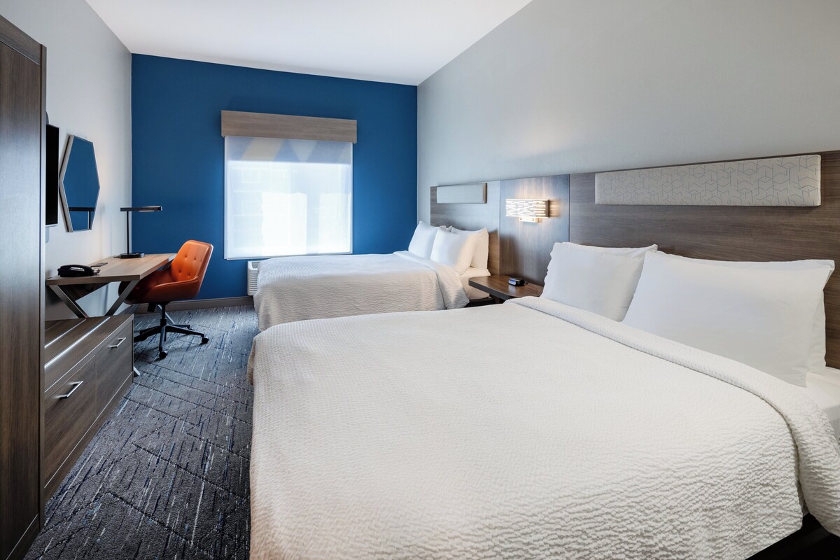 A hotel room with two beds, a desk with a chair, a dresser, and a window with blinds.
