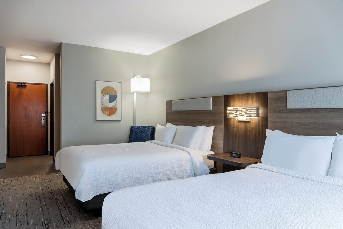 A hotel room with two beds, a bedside table, a lamp, and a wall-mounted artwork.