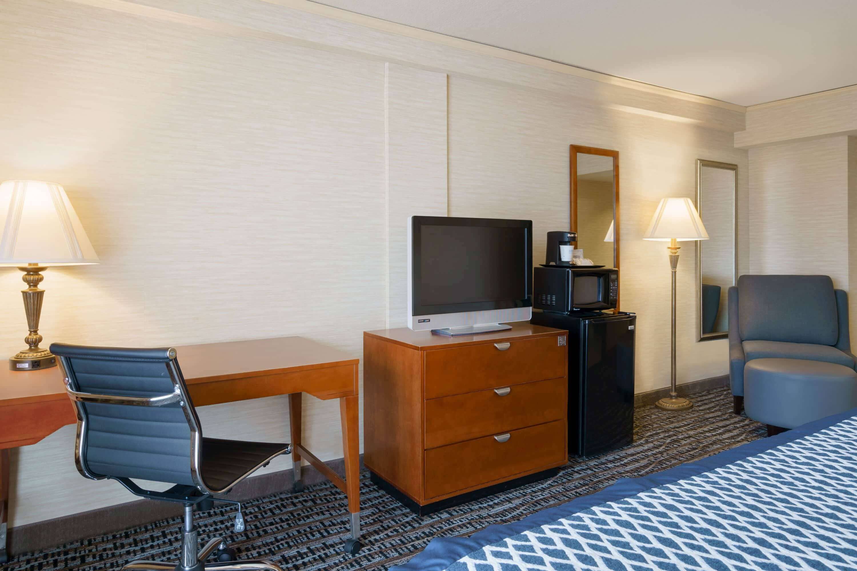 Quarto de hotel com uma cama, uma mesa com cadeira, televisão, cafeteira, luminária e cômoda.