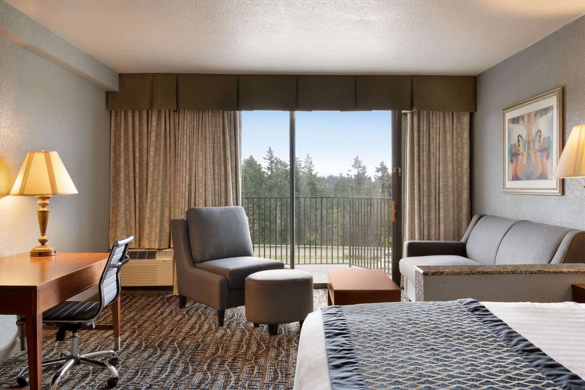 A hotel room with a large bed, a desk with a lamp, two armchairs, a small table, and a view of trees through the window.