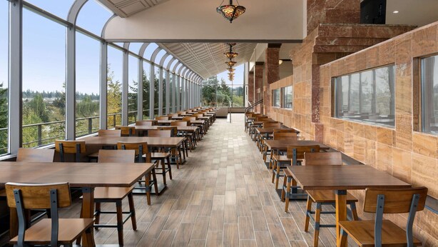 Un comedor espacioso con mesas y sillas largas, ventanas amplias que ofrecen vistas al exterior y lámparas modernas.