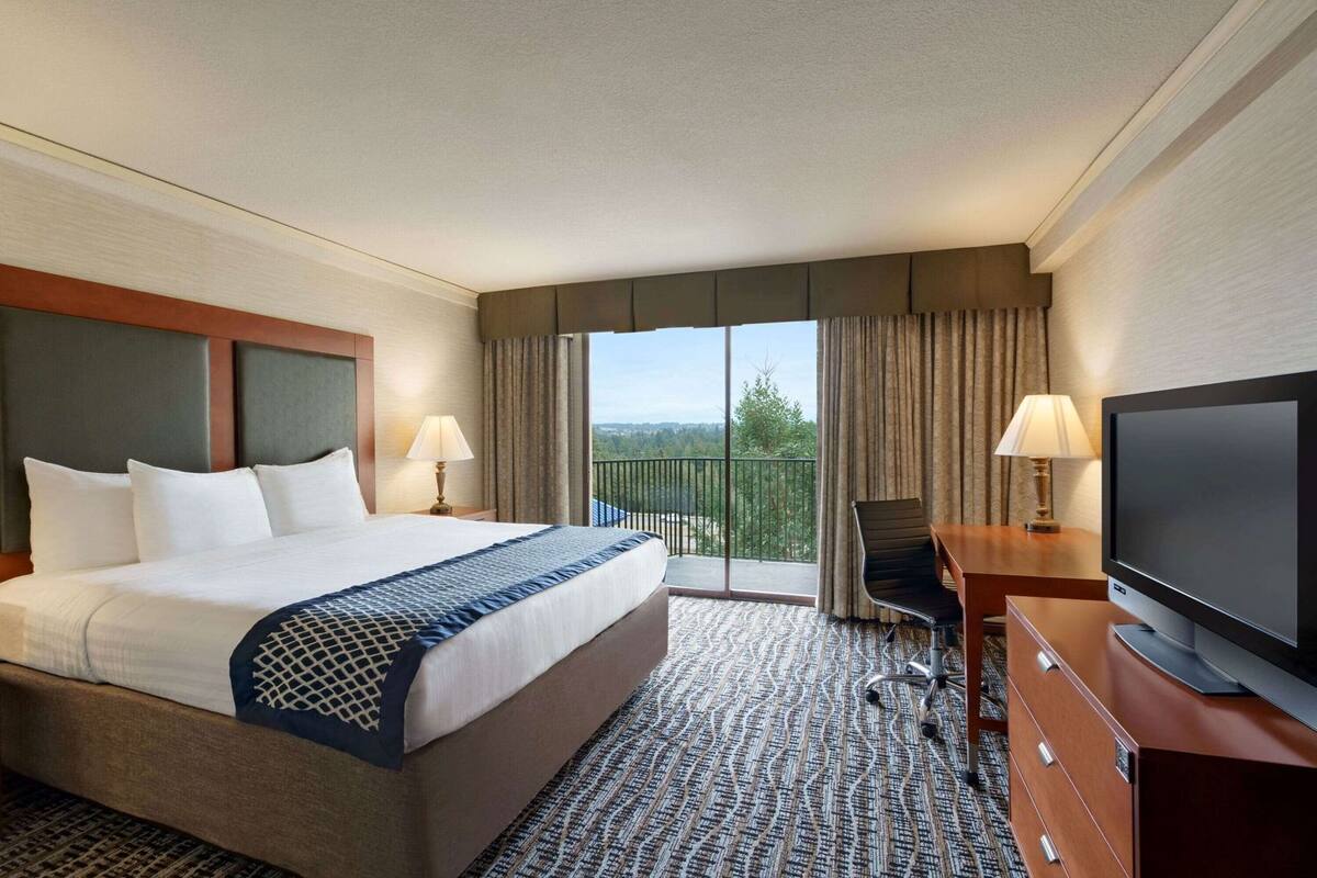 A hotel room with a large bed, a desk with a TV, a chair, and a balcony with a view.