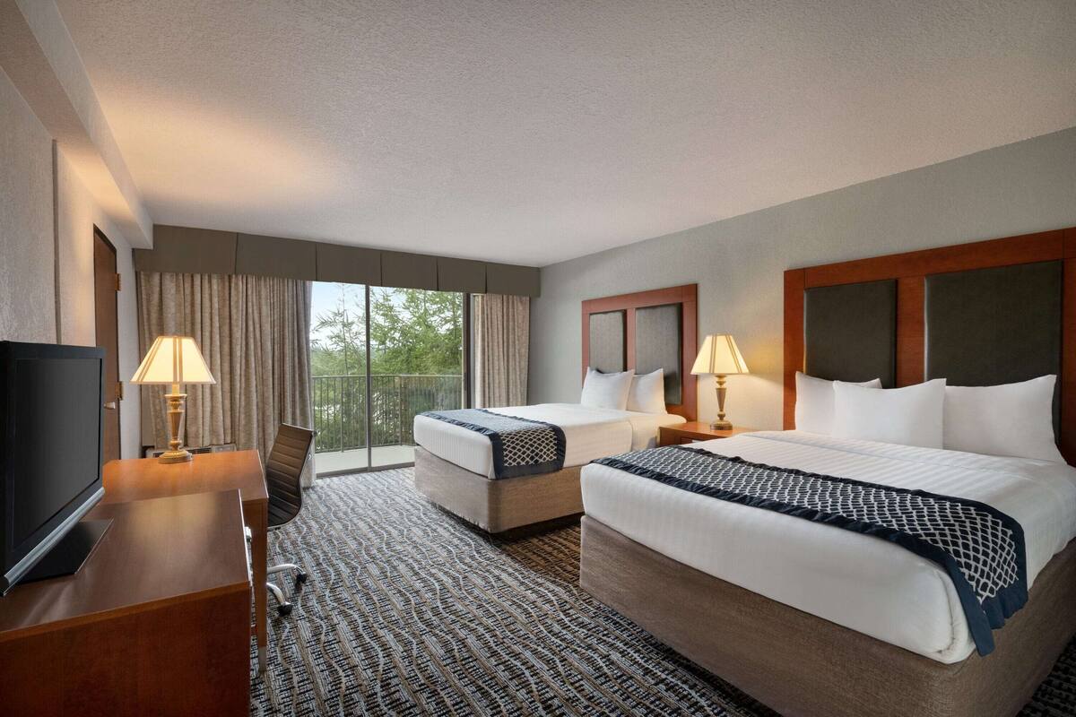 A hotel room with two beds, a desk with a lamp, a TV, and a large window with curtains.
