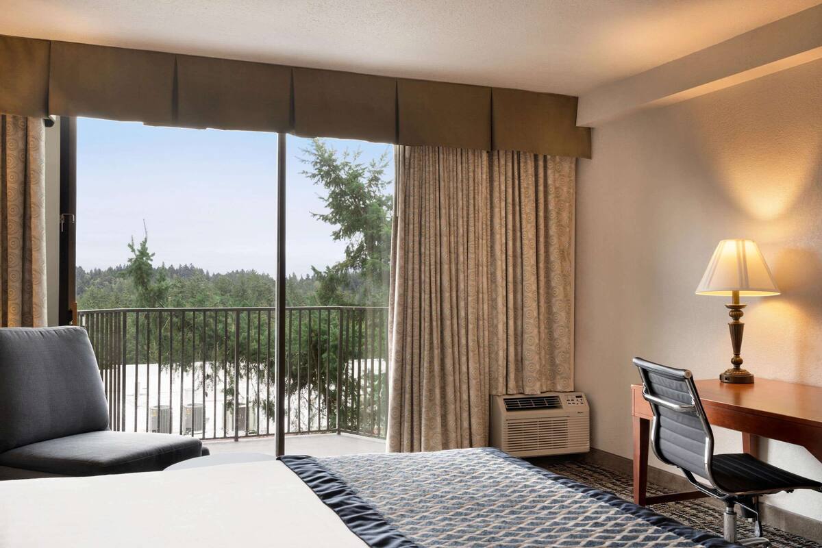 A hotel room with a bed, a chair, a desk, a lamp, and a view of a balcony with a railing and trees.