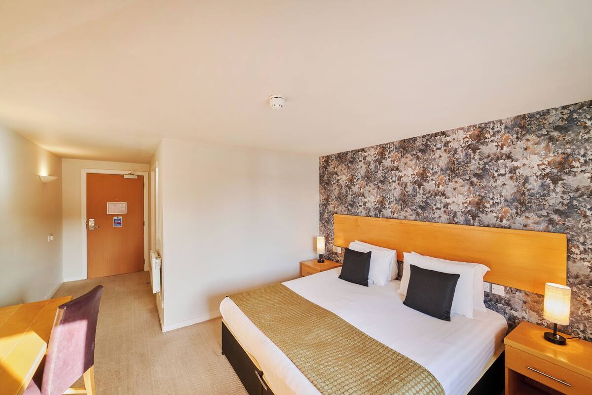 A hotel room with a large bed, a wooden headboard, a nightstand with a lamp, a desk with a chair, and a door leading to another room.