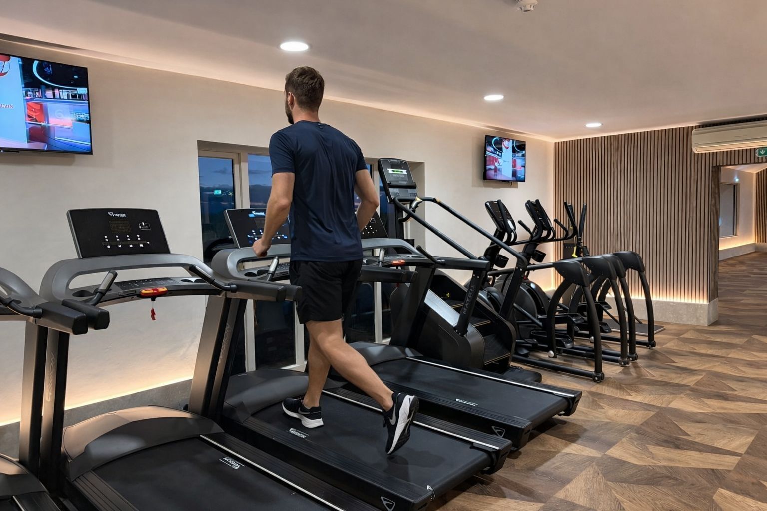 A gym with treadmills, a TV screen, and a wooden wall.