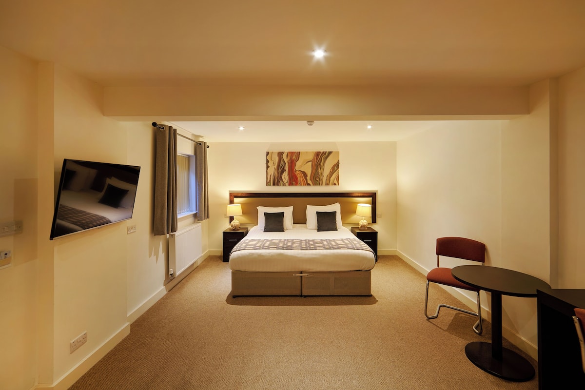 A hotel room with a large bed, a flat-screen TV mounted on the wall, a desk with a chair, and a painting above the bed.
