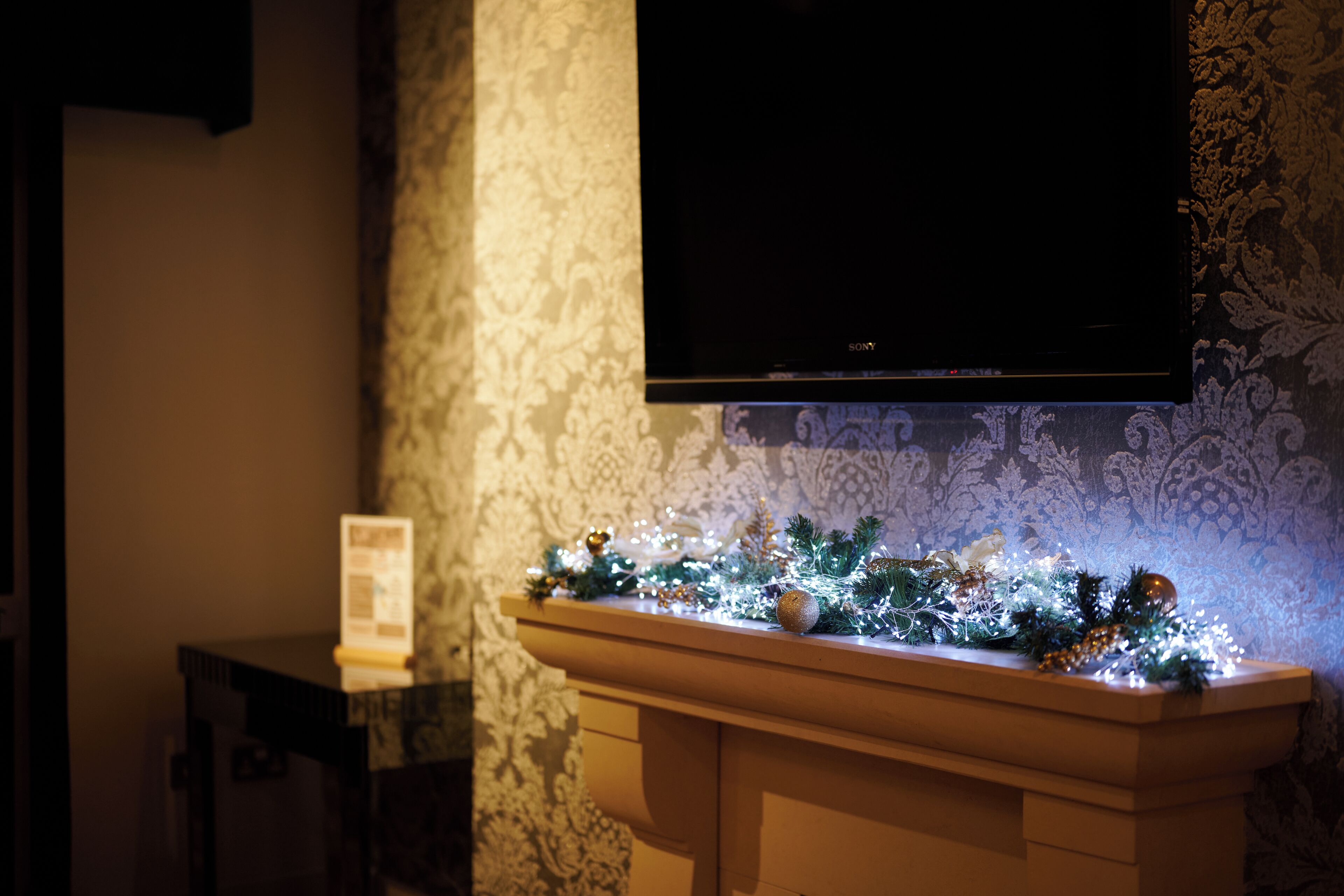 A fireplace with Christmas decorations, a television mounted on the wall, and a patterned wallpaper.