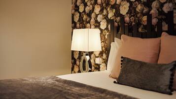 A neatly made bed with a patterned bedspread, a bedside lamp, and a floral wallpaper backdrop.