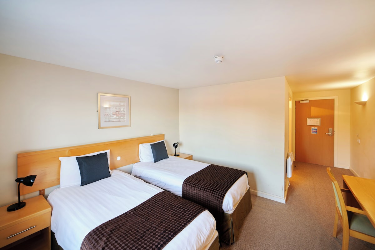A hotel room with two beds, a desk, and a door leading to a hallway.