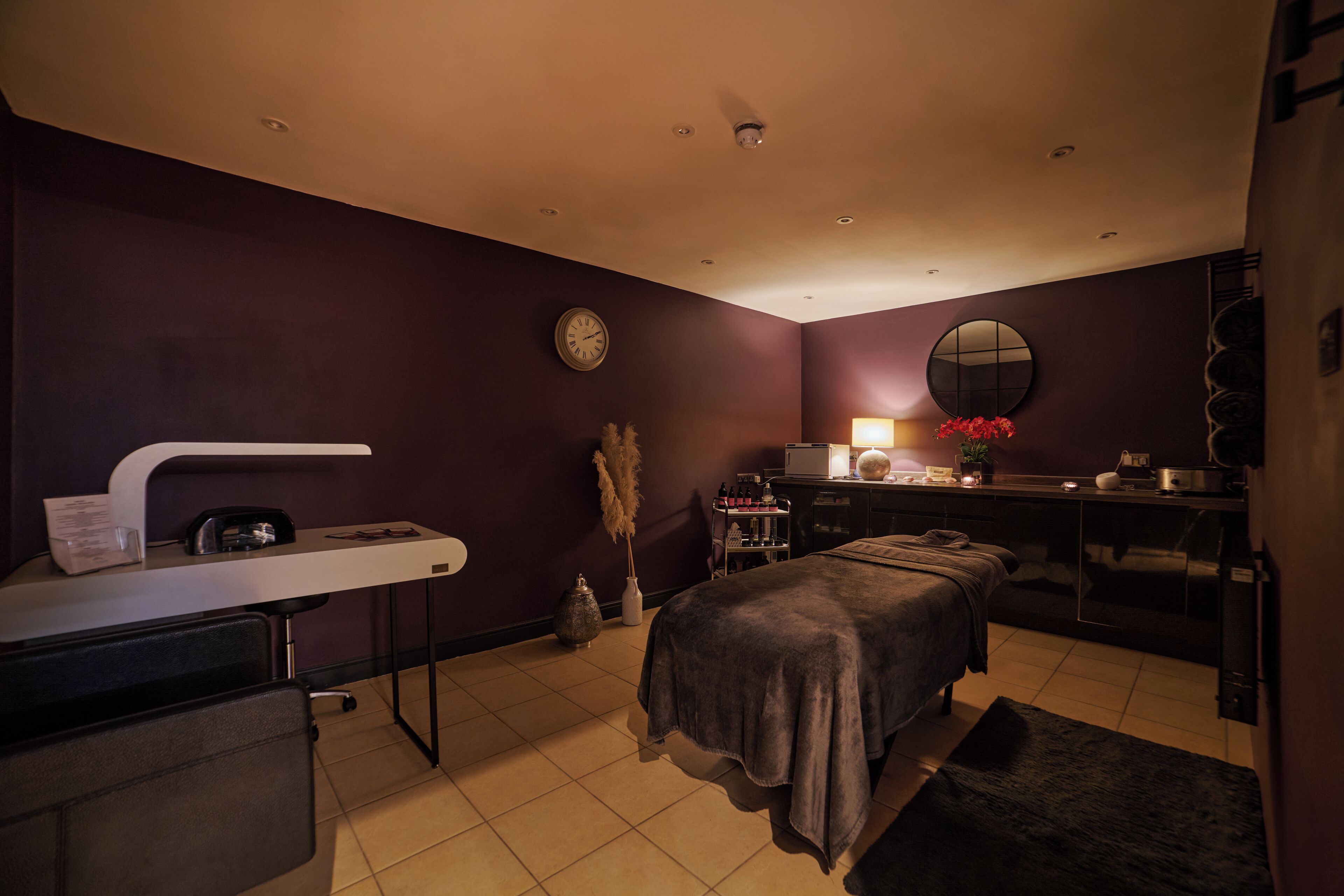 Un cuarto de spa con una camilla de masaje, un escritorio con lámpara, un reloj y un espejo en la pared.