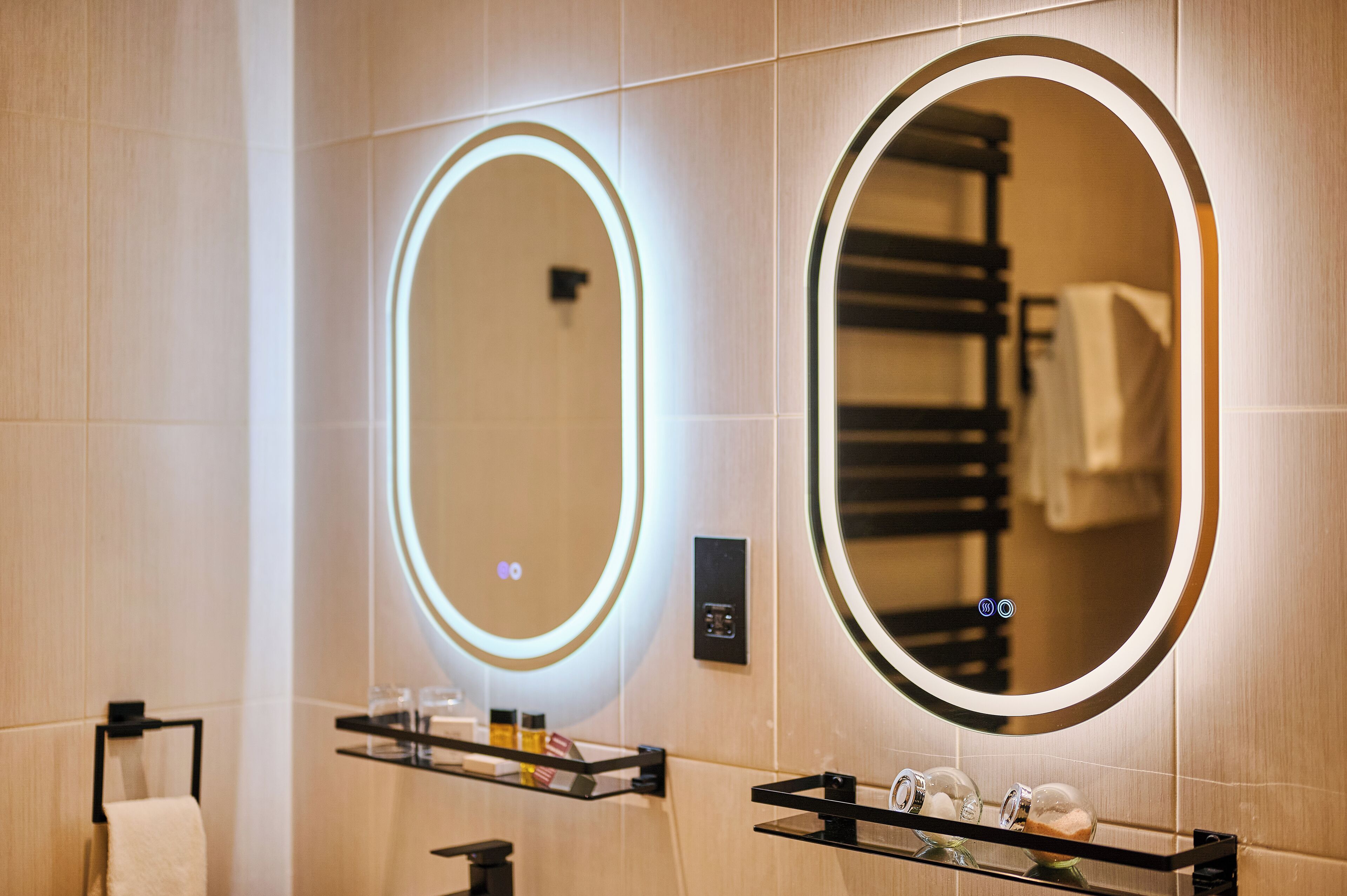 Un baño moderno con dos espejos iluminados, un perchero para toallas y un estante con artículos de aseo.
