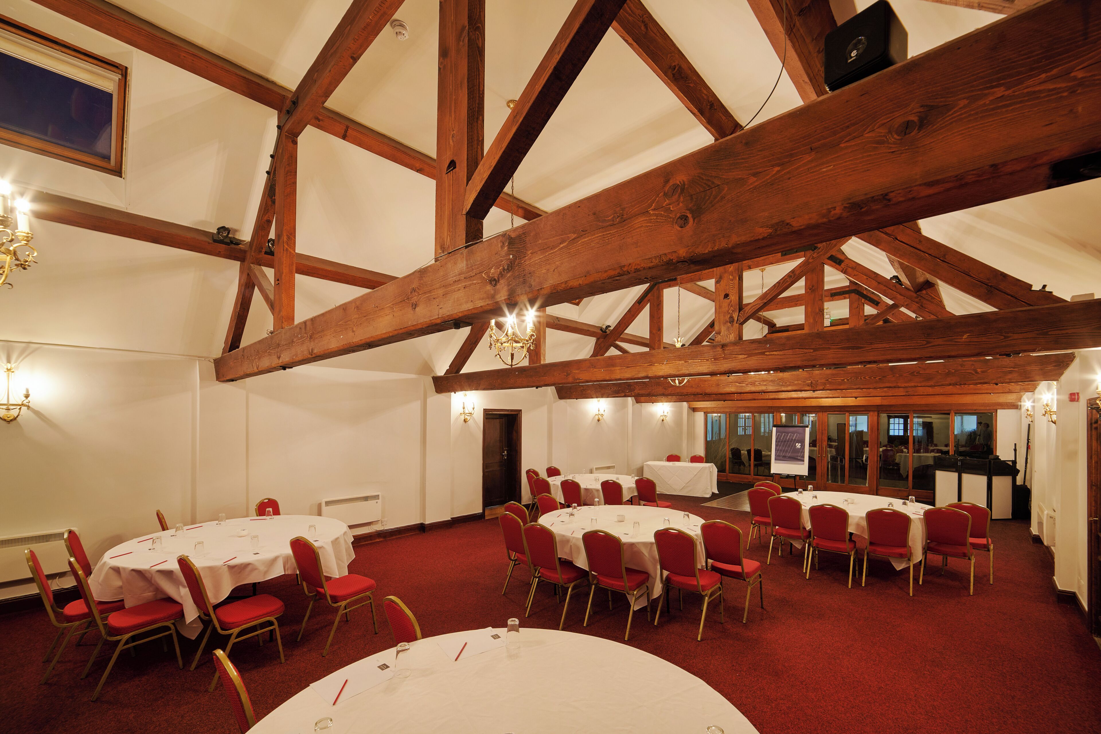 A conference room with multiple round tables and red chairs, wooden beams on the ceiling, and a white carpet.