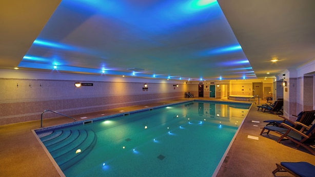 Une piscine intérieure avec un éclairage bleu, un mur carrelé et des chaises longues disposées le long du bord.