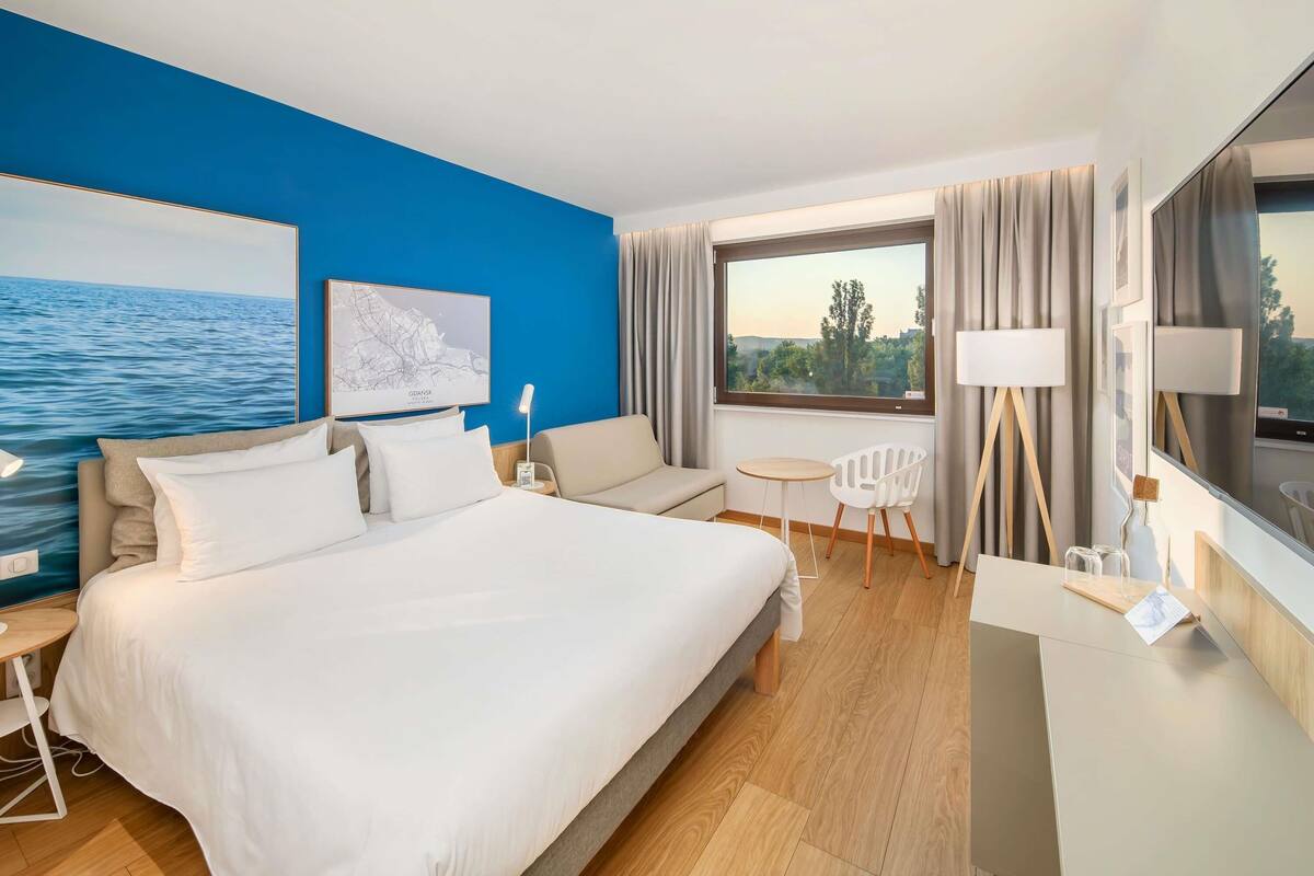 A hotel room with a large bed, a desk, a chair, a small table, a lamp, and a window with a view of trees and a body of water.