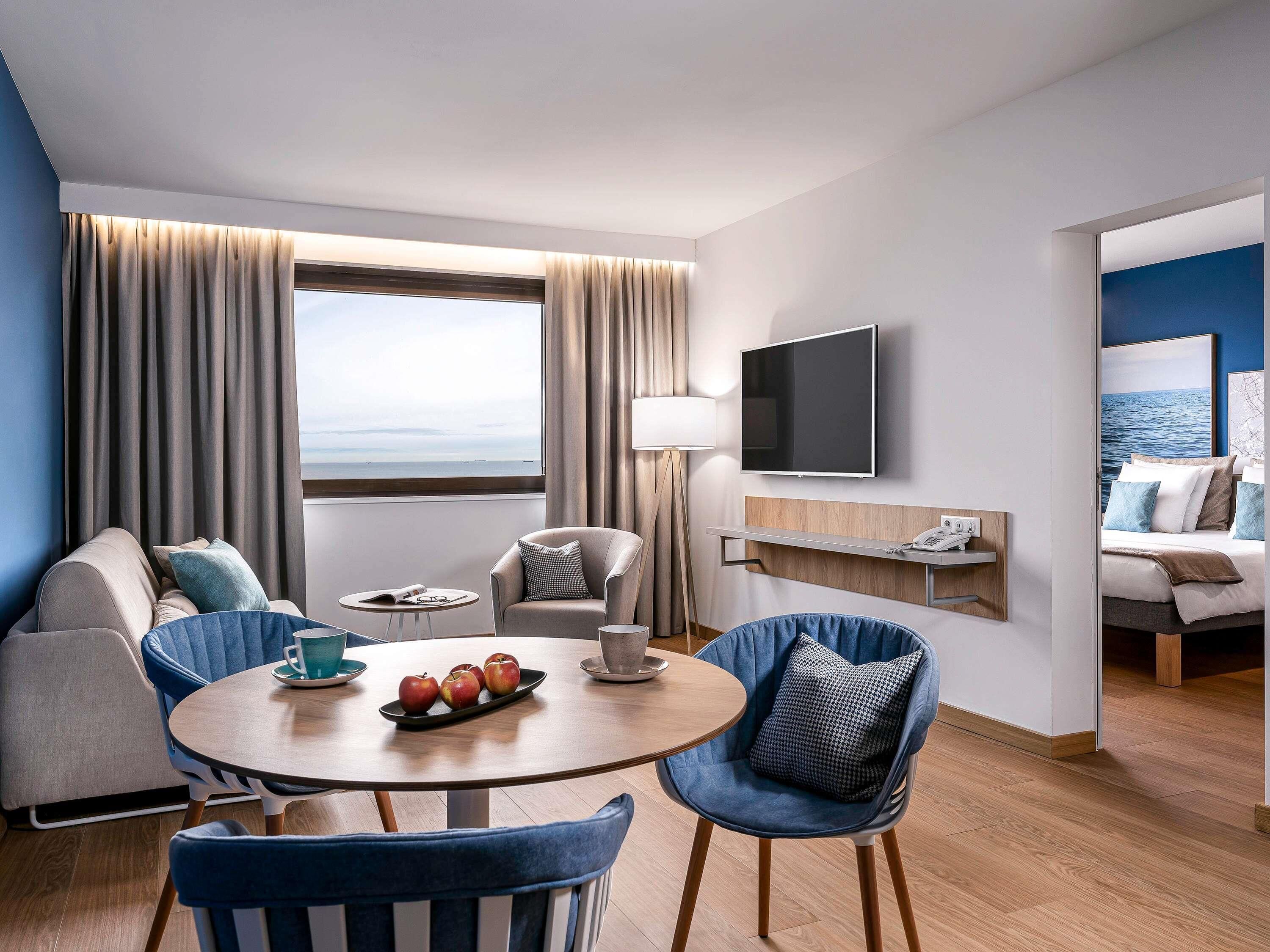 Et moderne hotelværelse med stort vindue, fladskærms-tv, sofa, spisebord med æbler og havudsigt.