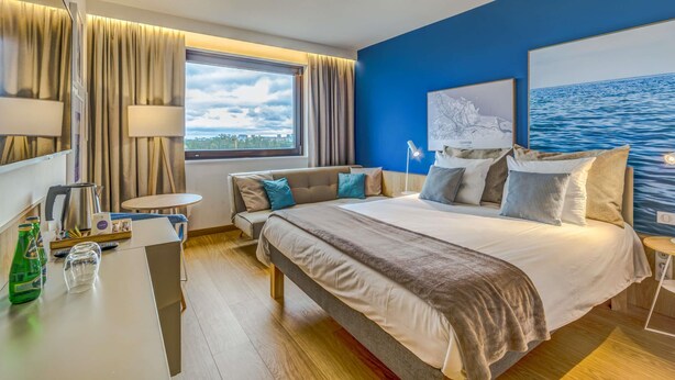 A modern hotel room with a large bed, a desk, a chair, a window with curtains, and a view of the sea. - Novotel Gdansk Marina (Gdansk)