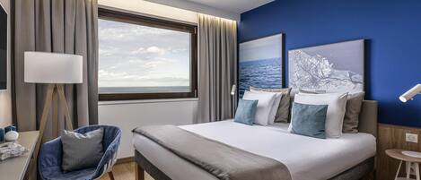 Une chambre d’hôtel avec un grand lit, un bureau avec une chaise, une lampe sur pied et une vue sur la mer.