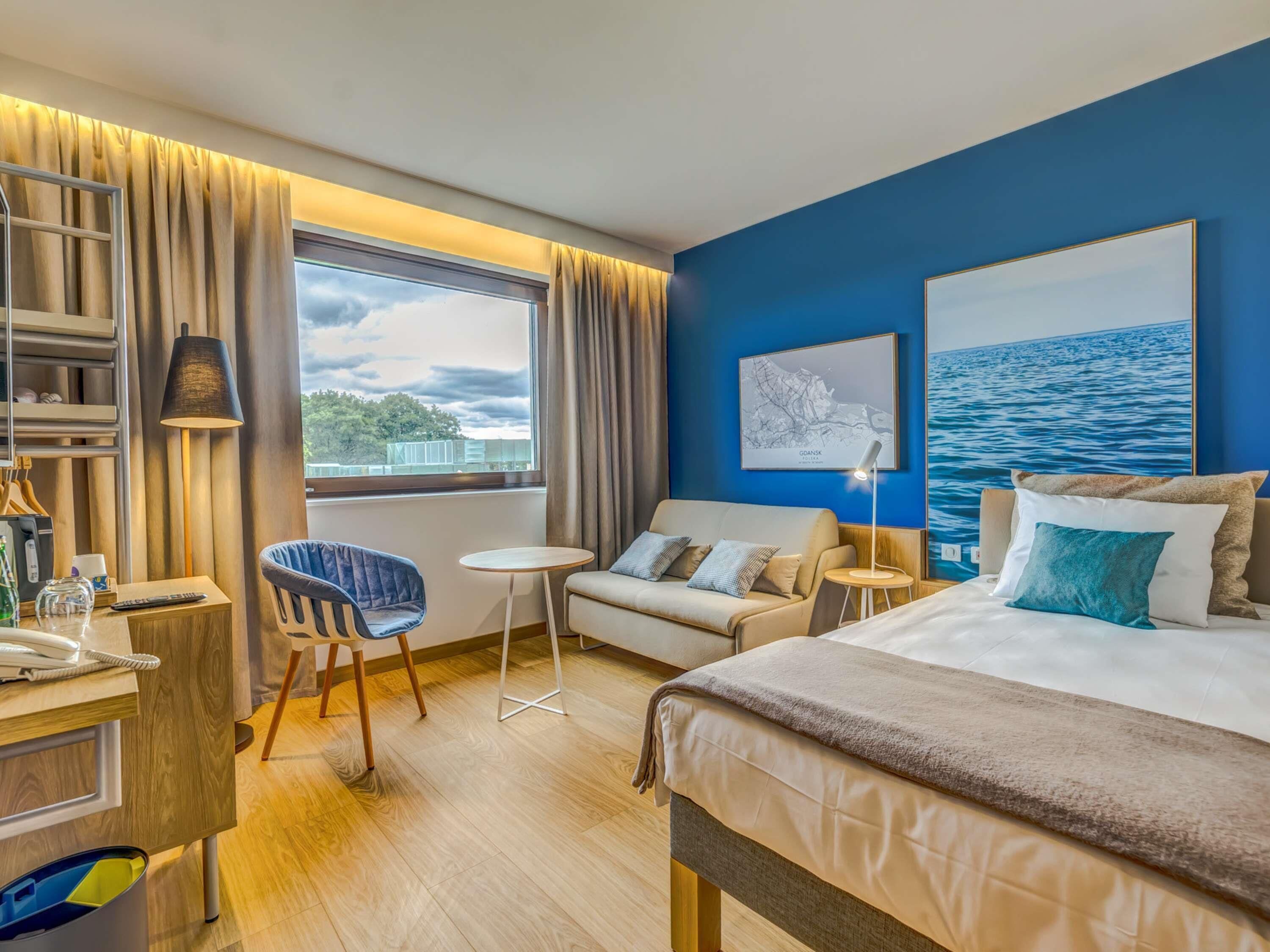 A hotel room with a bed, a sofa, a chair, a desk, a lamp, a window, and a wall with a map and a painting.
