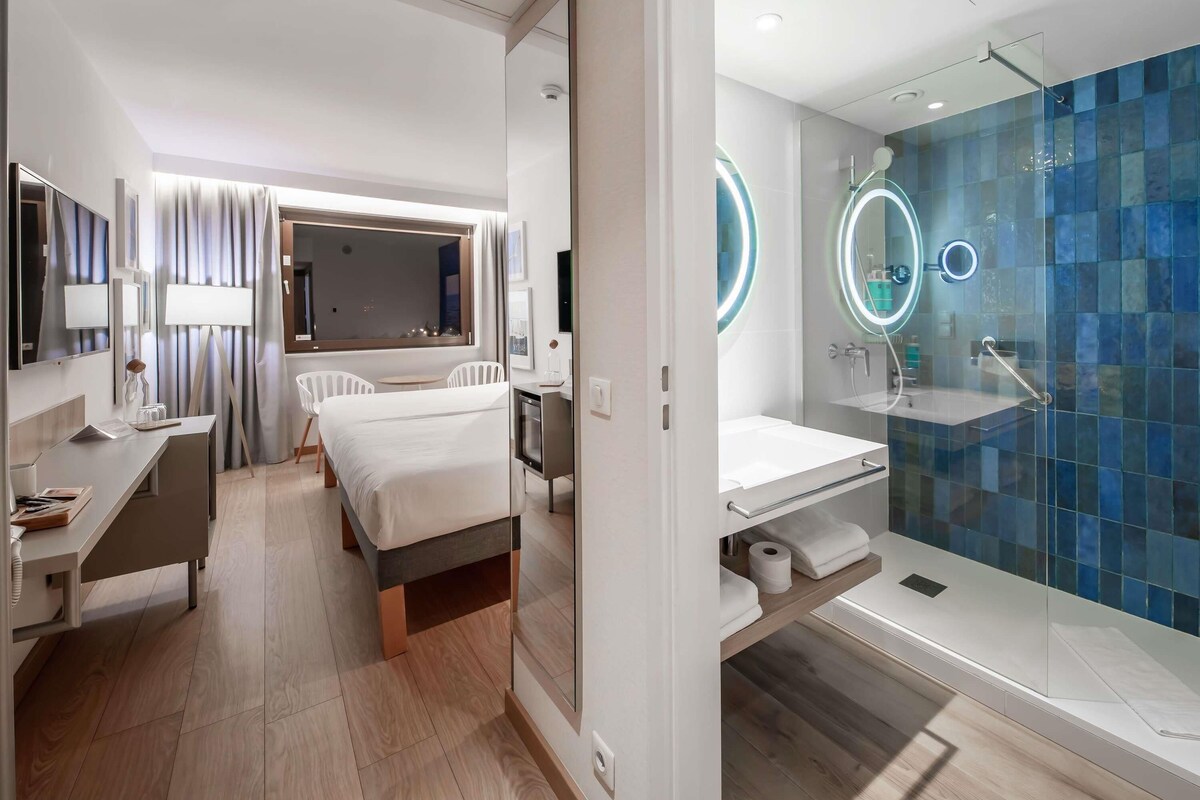 Moderni hotellihuone, jossa on sänky, työpöytä ja kylpyhuone lasiseinällä varustetulla suihkulla.
