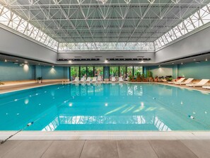 An indoor swimming pool with a high ceiling, clear glass panels, and lounge chairs along the sides.