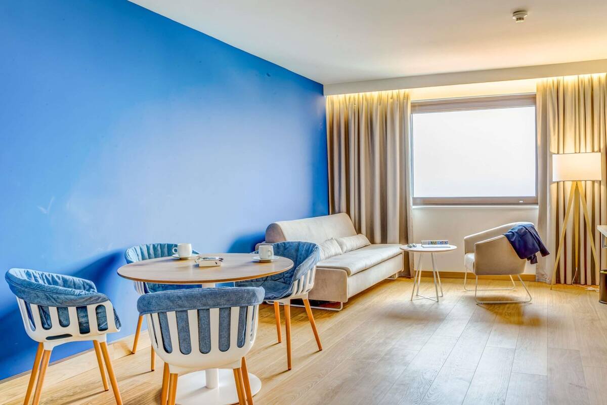 A modern hotel room with a blue accent wall, a round dining table, two blue armchairs, a white sofa, a small side table, a lamp, and a large window with curtains.