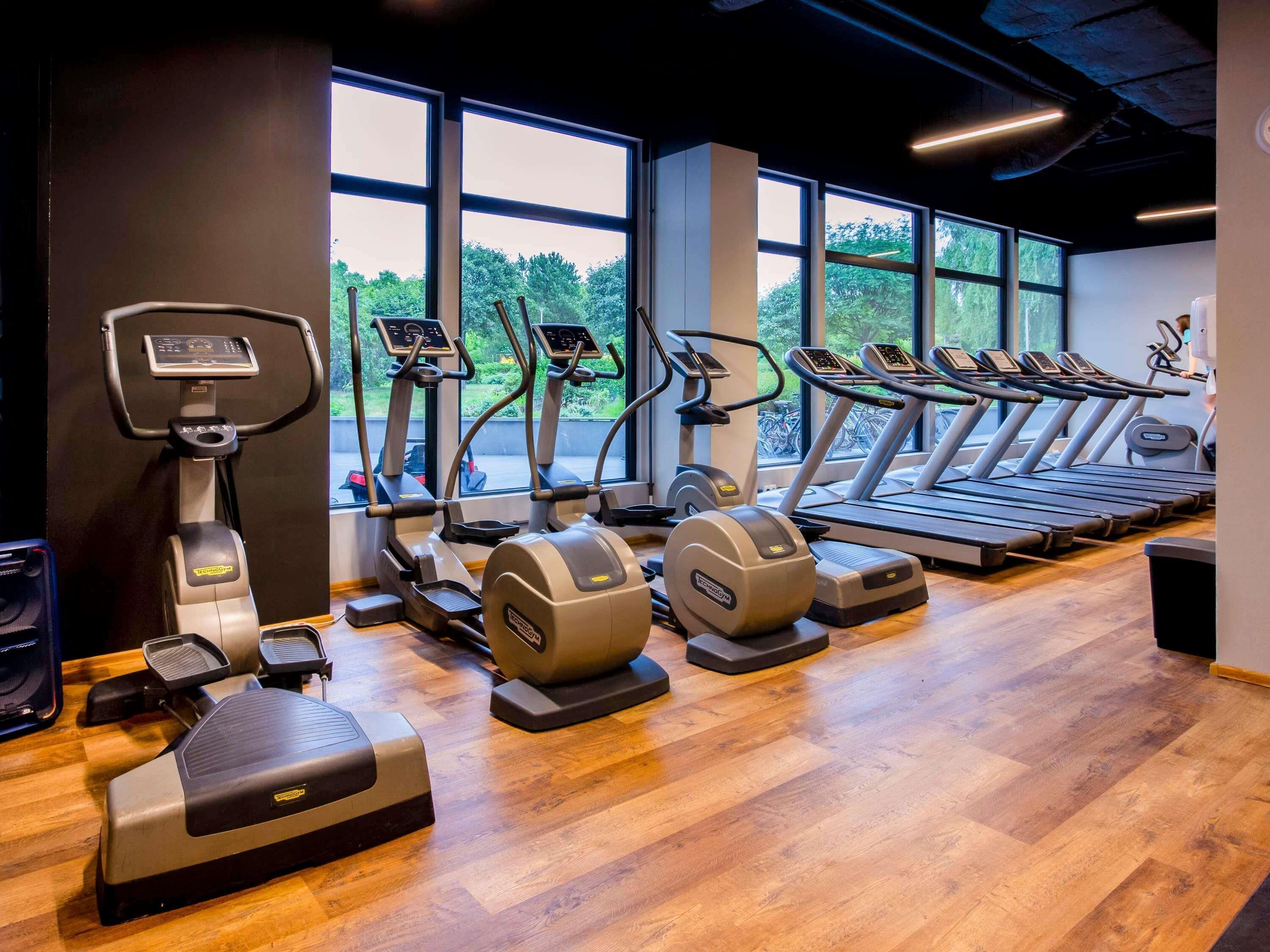 Un gimnasio con diversas máquinas de cardio, ventanas amplias y suelo de madera.