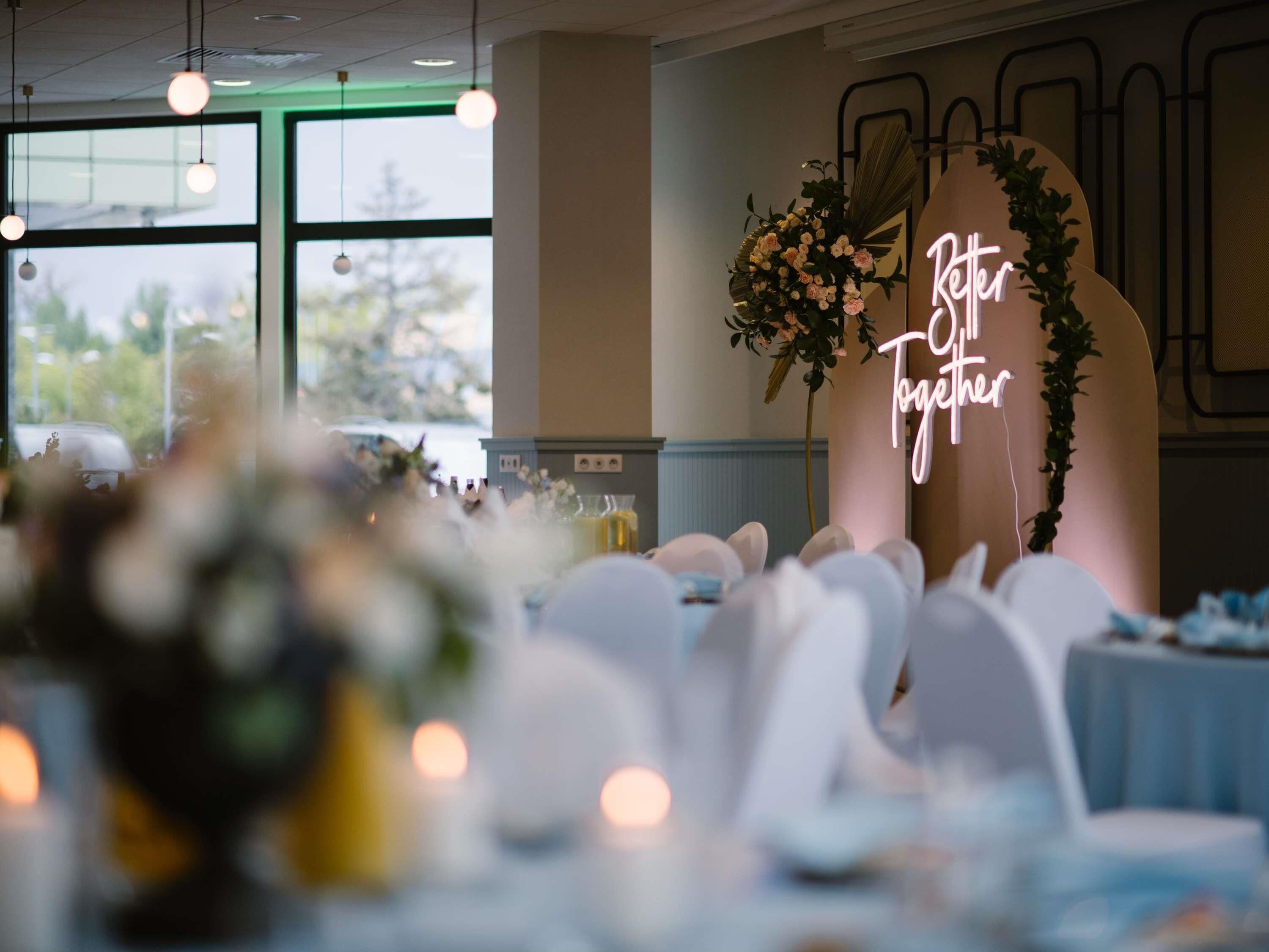 Salón de recepción de boda con un letrero de neón que dice 'Better Together', mesas con sillas blancas y un ventanal con vistas a árboles.