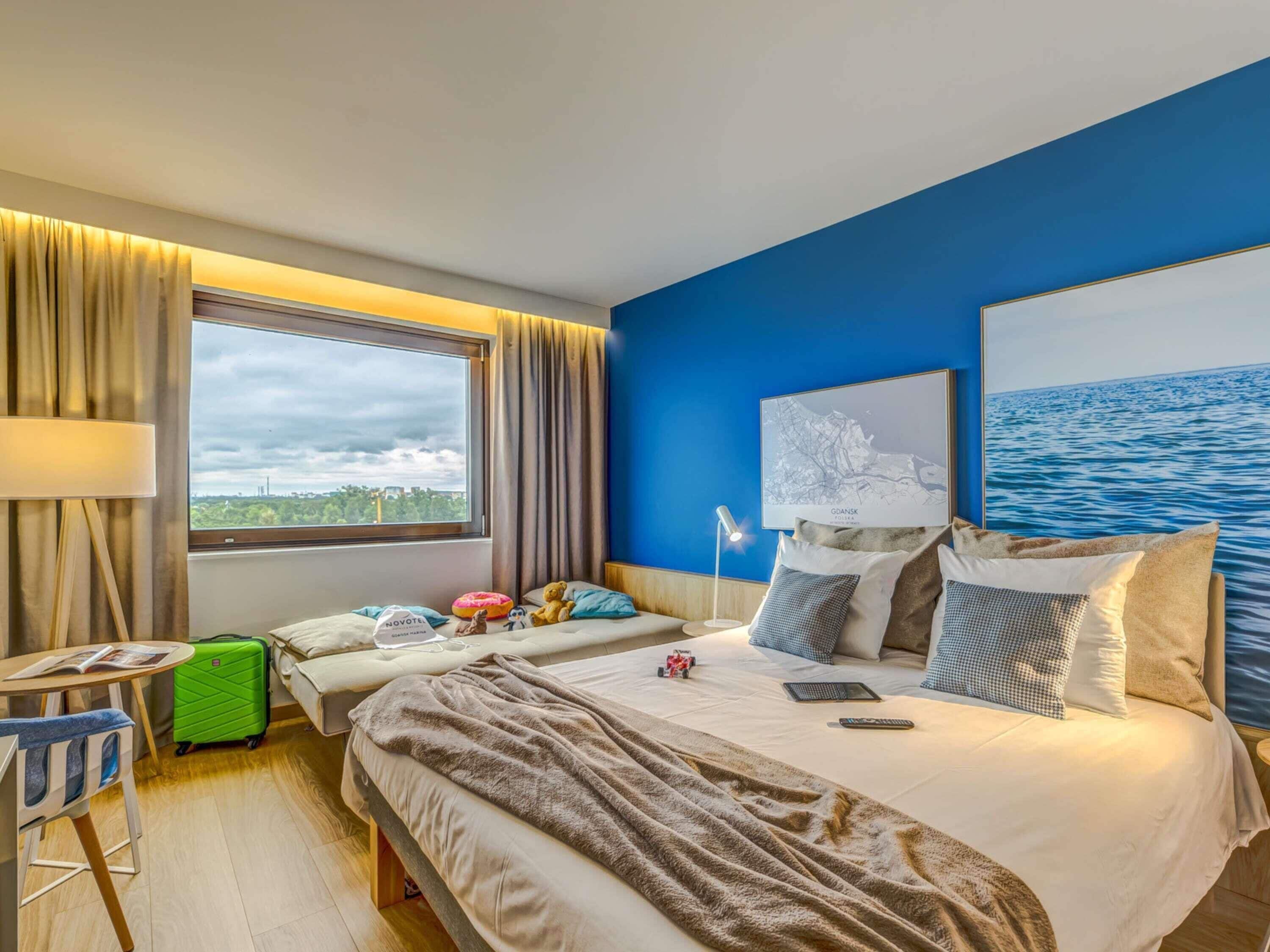 Habitación de hotel con cama, escritorio, silla, maleta y una ventana grande con vistas.