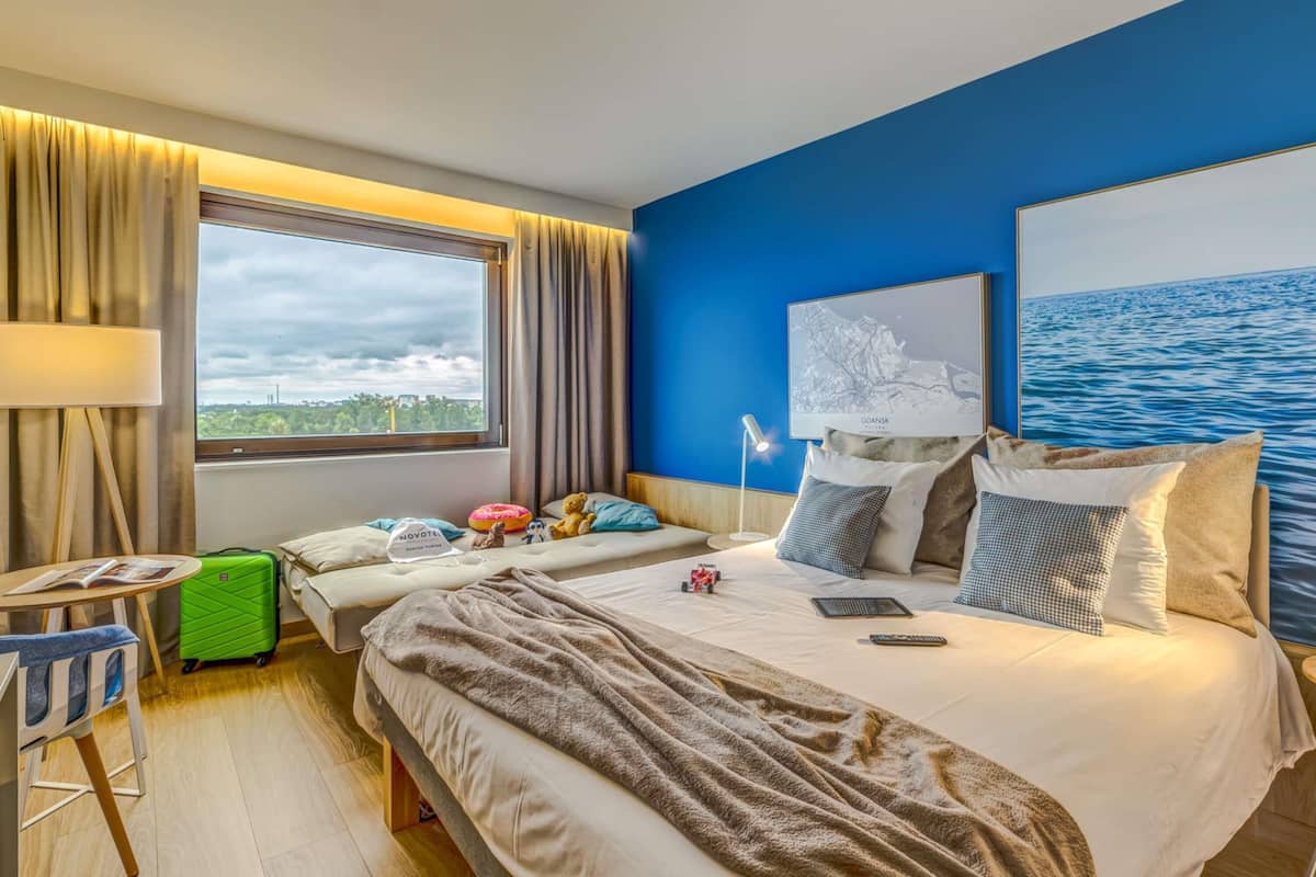 A hotel room with a bed, a desk, a chair, a suitcase, and a large window with a view.