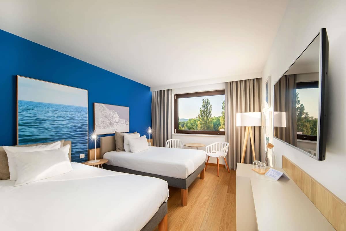 A hotel room with two beds, a desk, a chair, a TV, and a window with a view of trees and hills.