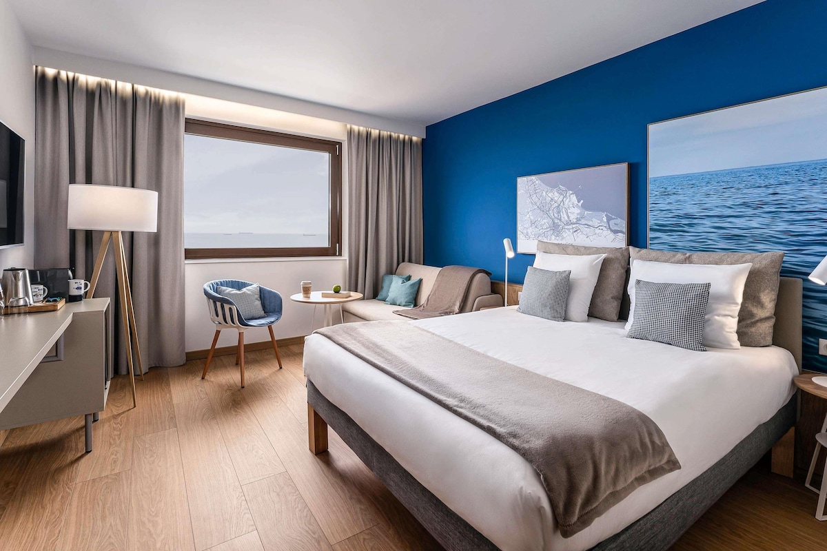 A hotel room with a bed, a desk, a chair, a small table, and a view of the sea.