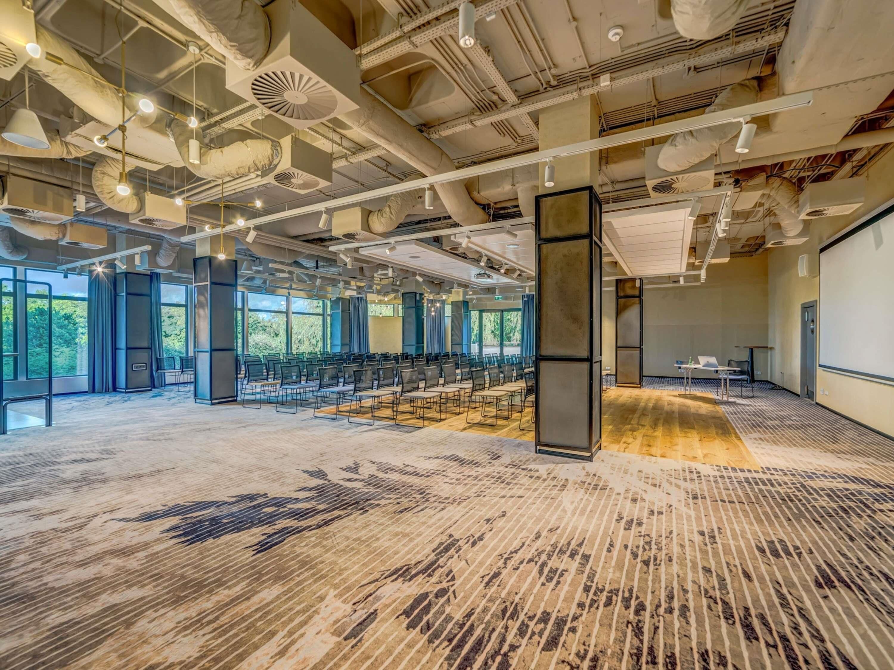 A spacious conference room with rows of chairs, large windows, and exposed ceiling pipes.
