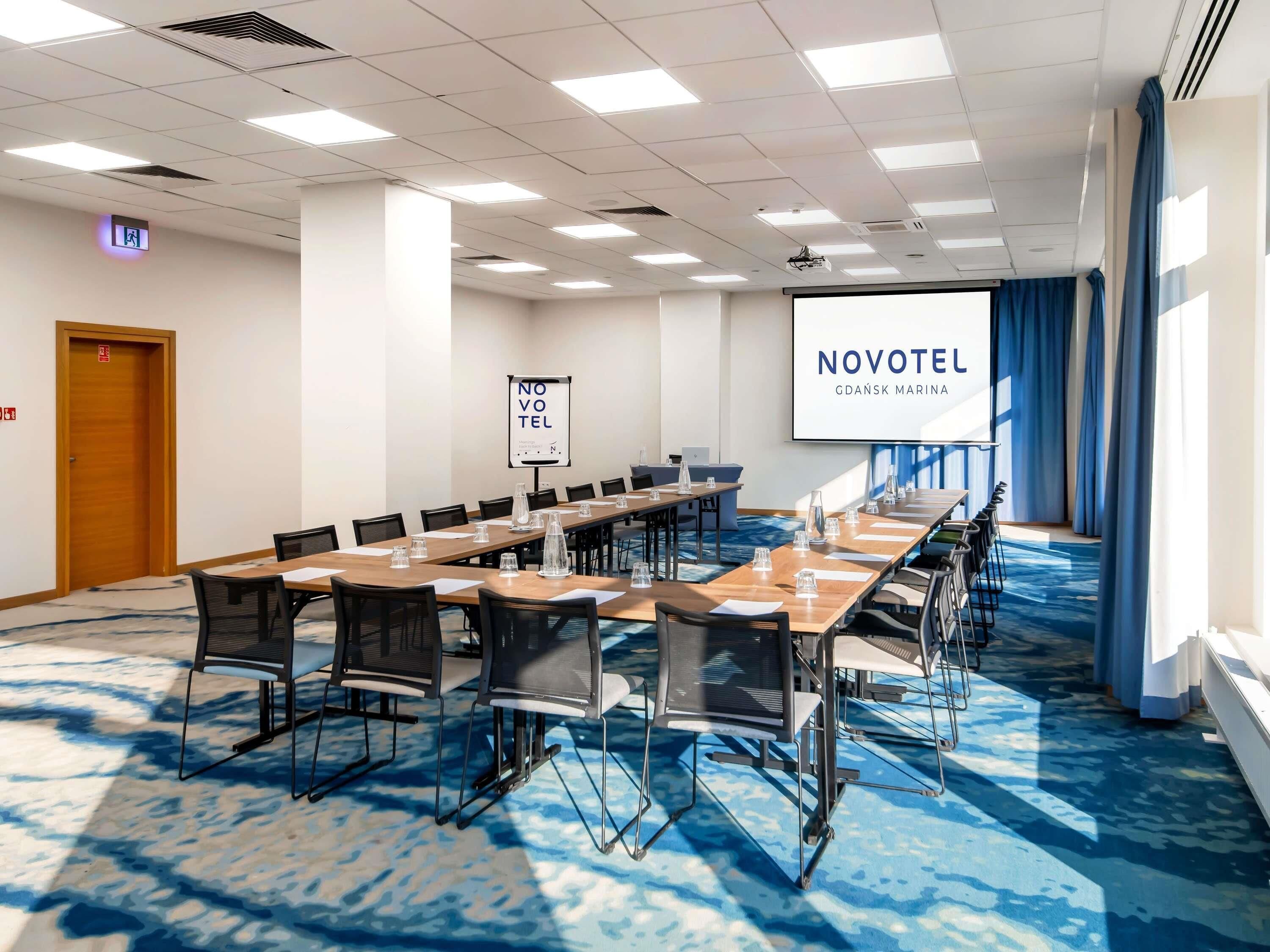 A conference room with a long table, chairs, a projector screen, and a banner reading 'NOVOTEL GDAĽSK MARINA'.