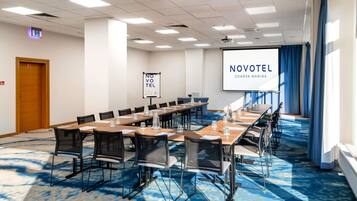 A conference room with a long table, chairs, a projector screen, and a banner reading 'NOVOTEL GDAĽSK MARINA'.