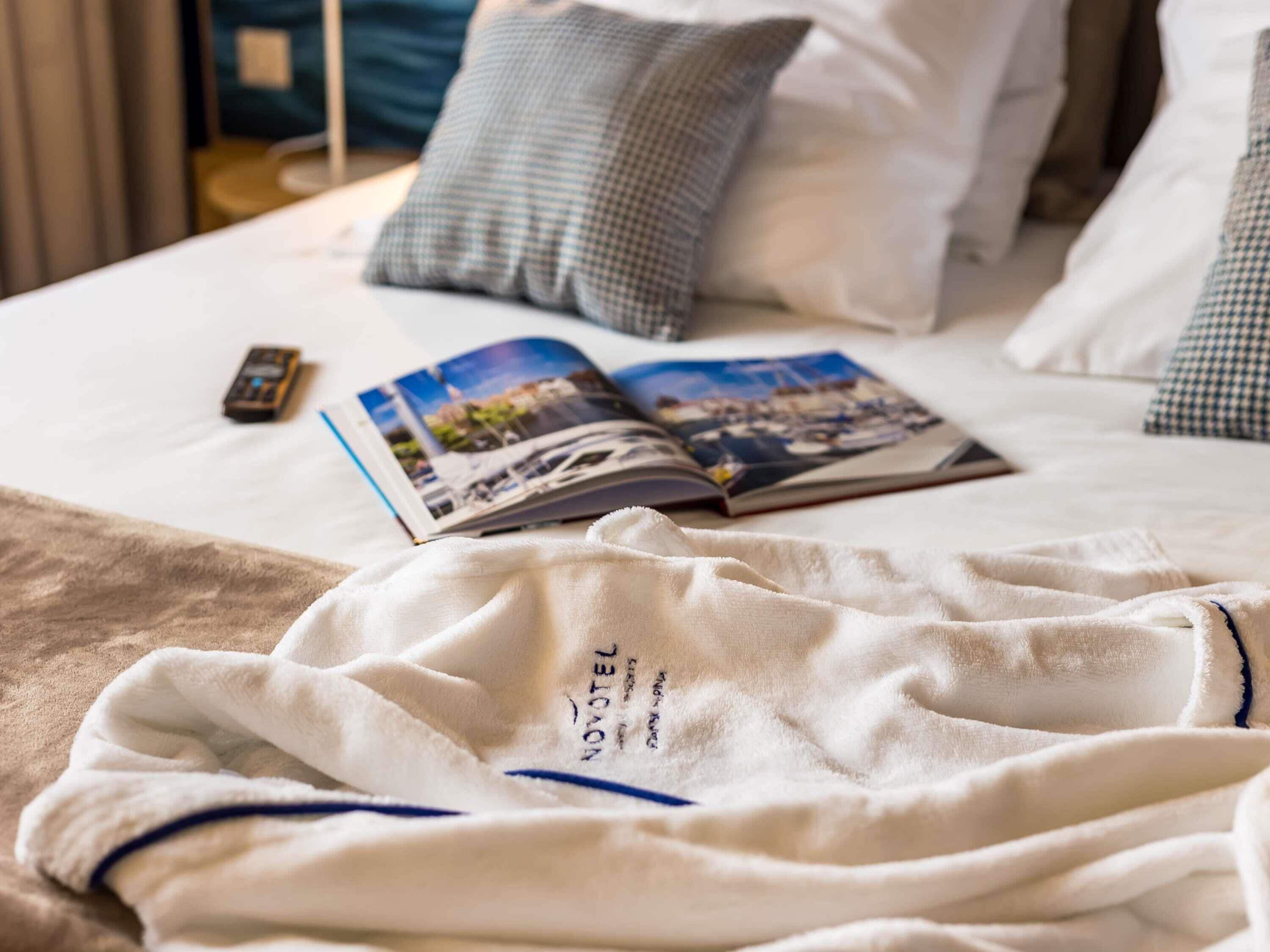 Ein ordentlich bezogenes Bett mit einer Zeitschrift, einer Fernbedienung und einem weißen Bademantel, auf dem "NOVOTEL" zu sehen ist.