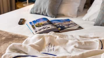 A neatly made bed with a magazine, a remote control, and a white robe with "NOVOTEL" visible.
