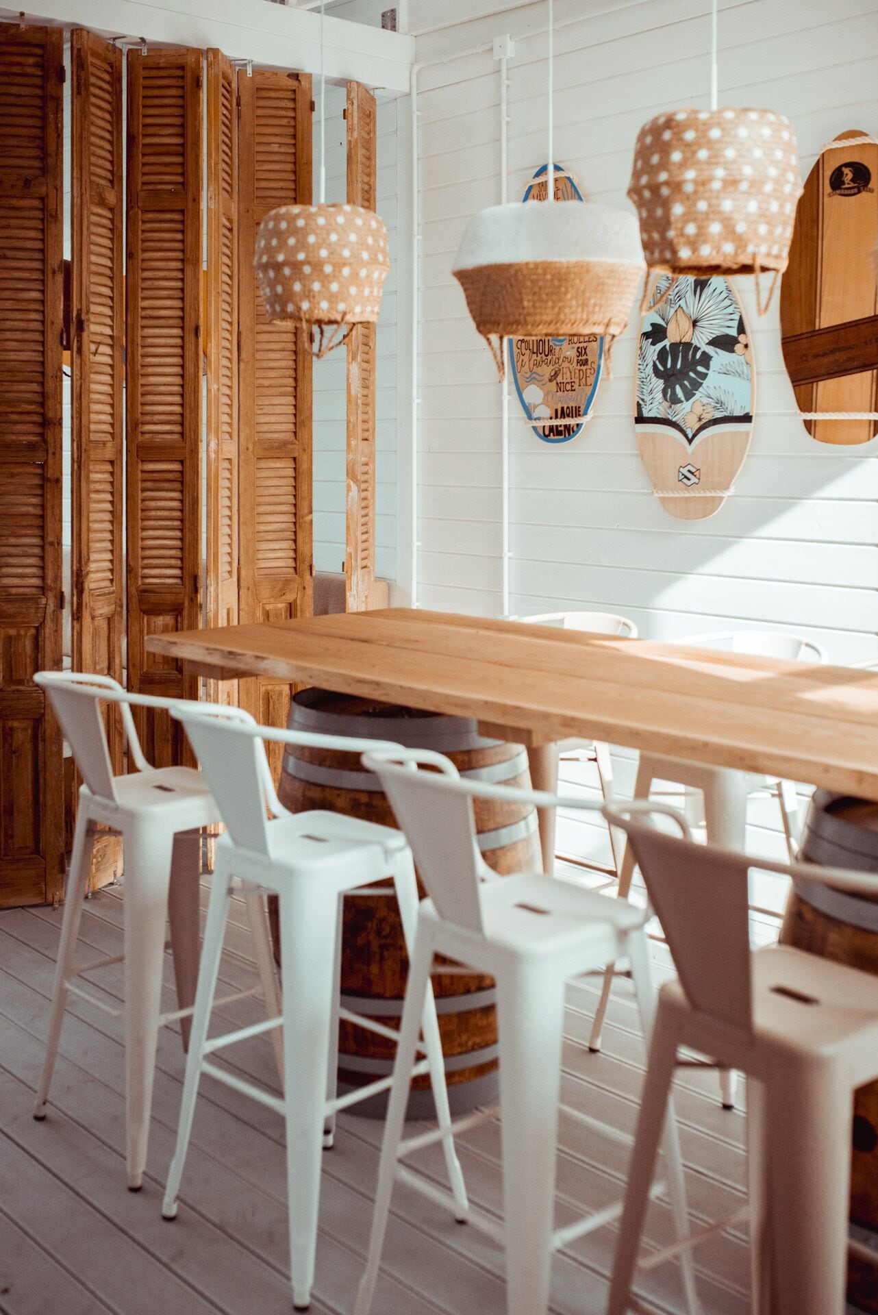 Une table à manger en bois, entourée de chaises blanches, un mur en bois avec des volets et un luminaire suspendu.