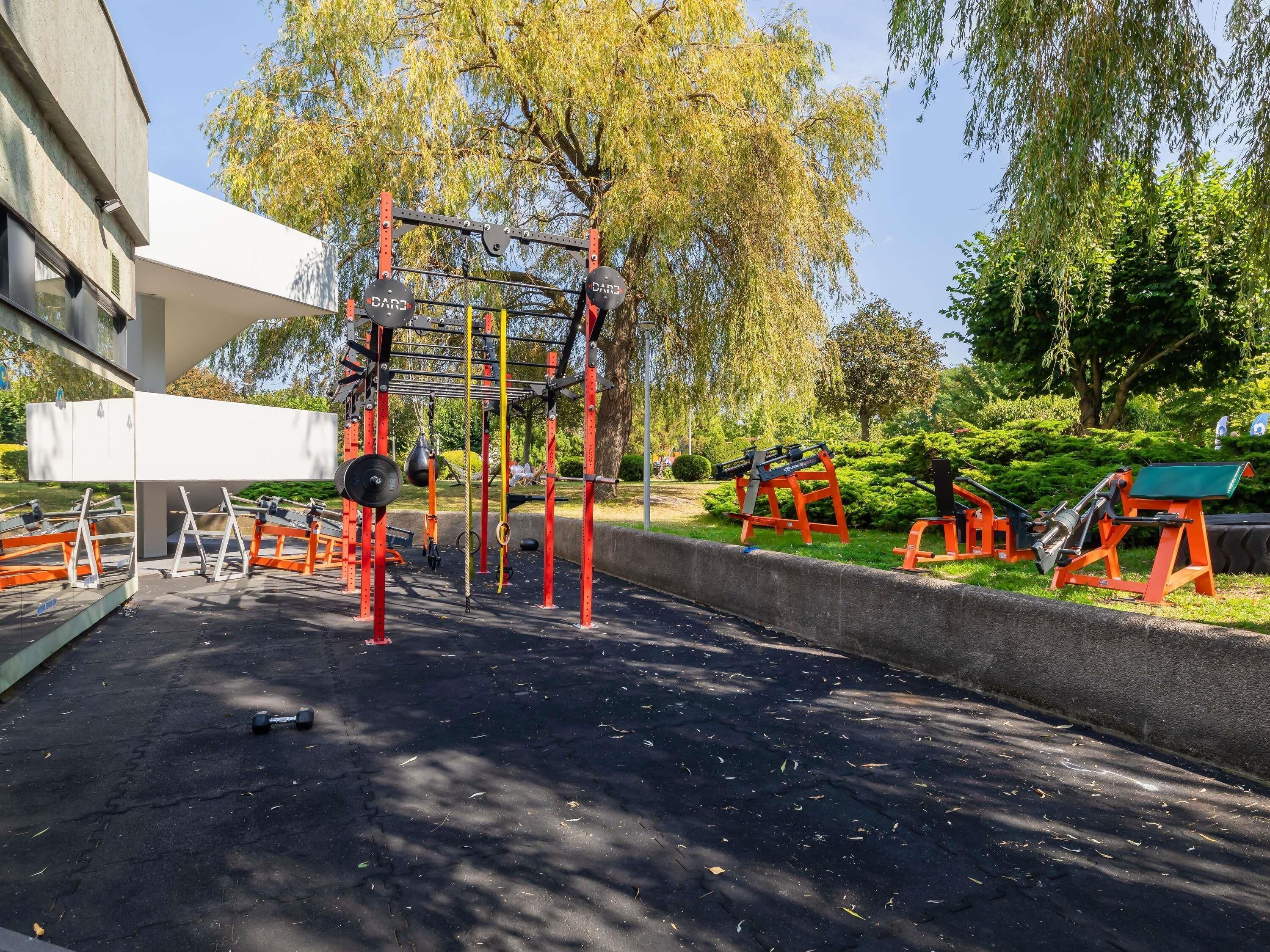 An outdoor gym with various exercise equipment, including pull-up bars and weight machines, set against a backdrop of trees and a building.