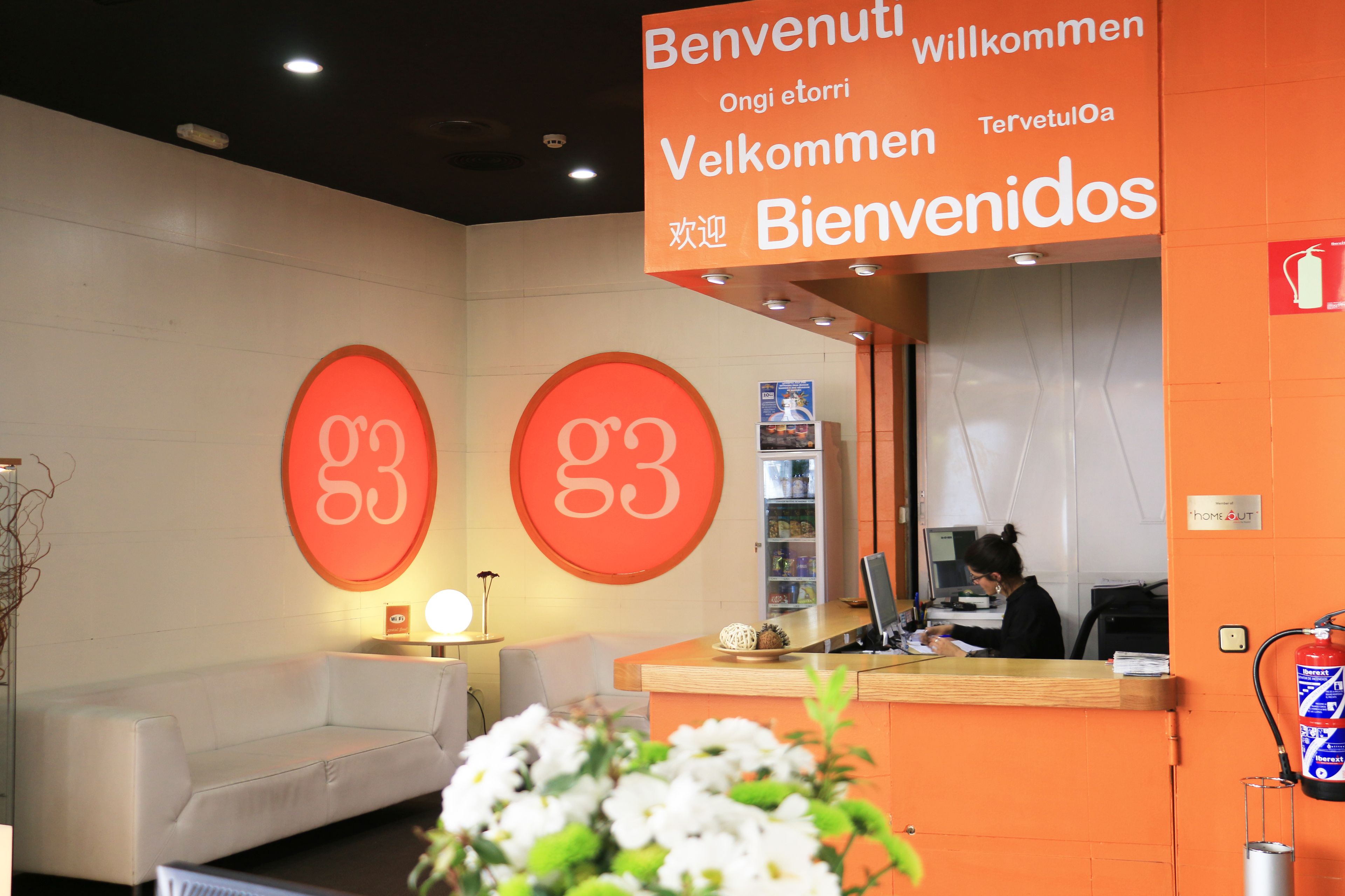 Un espace d’accueil avec une grande enseigne orange sur laquelle est écrit le mot « Bienvenue » dans plusieurs langues, un comptoir d’accueil, un canapé blanc et un extincteur fixé au mur.