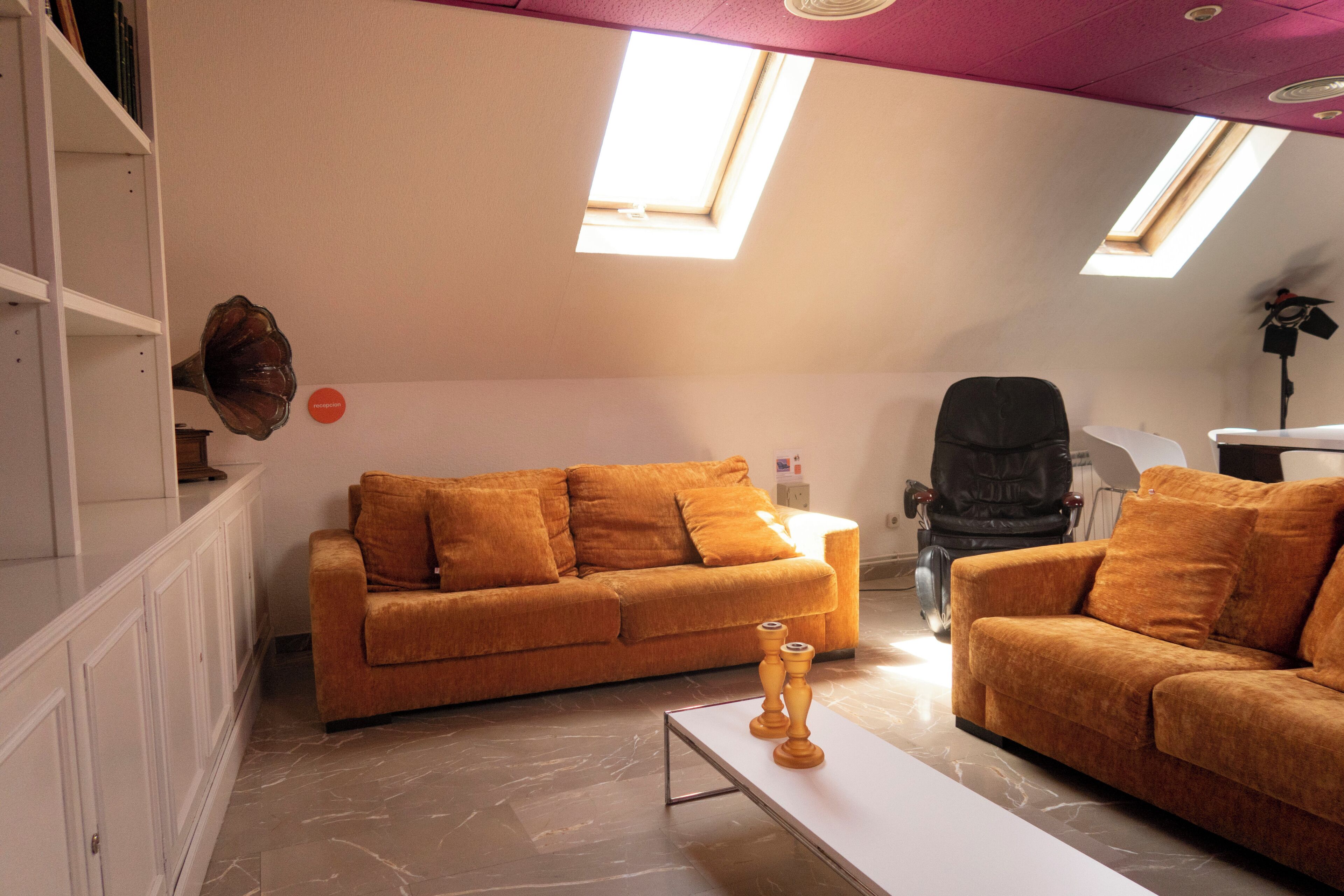 Una sala moderna con un sofá marrón, una mesa de centro blanca y una silla de escritorio negra. Hay tragaluces en el techo y un armario blanco con estantes.