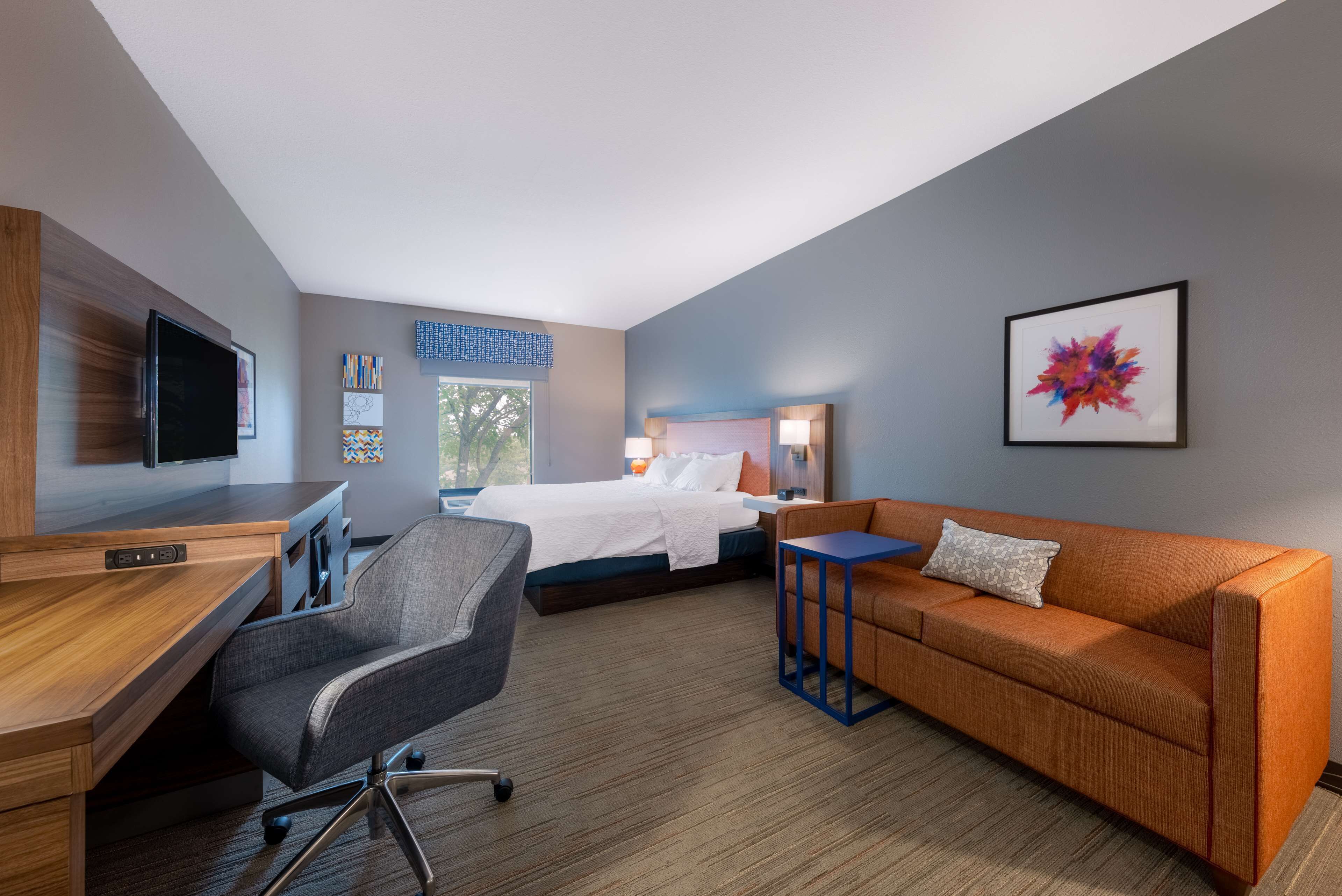 A hotel room with a bed, a desk, a chair, a sofa, a TV, and artwork on the walls.