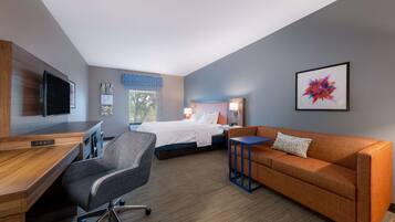 A hotel room with a bed, a desk, a chair, a sofa, a TV, and artwork on the walls.